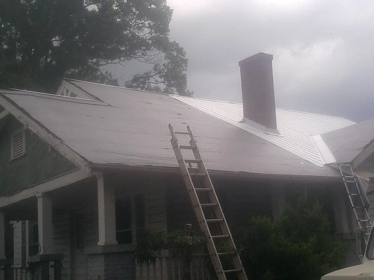Ladders against a metal roof on an older home with a brick chimney in Cost