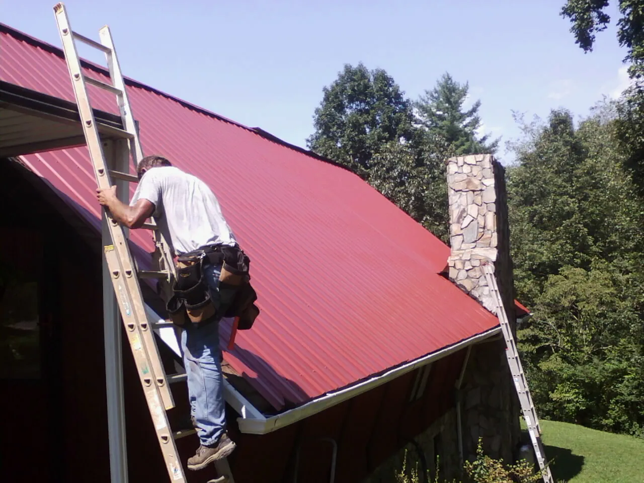 Roofer on a ladder beside a red metal roof and stone chimney — Soffit & Fascia Repair by Roofing Brothers in Cost